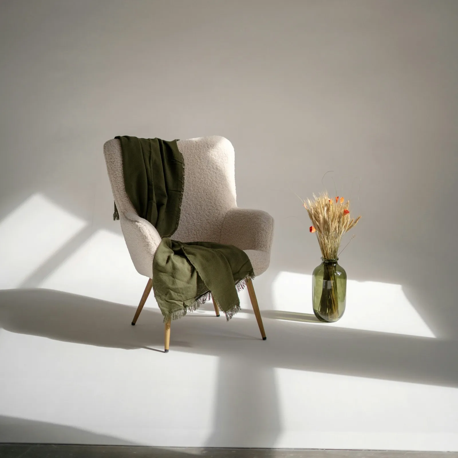 A cream armchair with an olive-green throw beside a vase of dried wheat